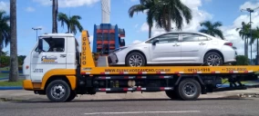 Guincho plataforma transportando um carro sedã branco em Belém, com árvores ao fundo e céu claro.