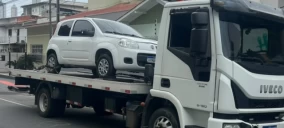Guincho plataforma transportando um carro branco, modelo Fiat, em Balneário Camboriú, estacionado na rua.