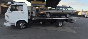 Guincho plataforma branco estacionado em posto de gasolina, transportando um veículo modelo station wagon cinza.