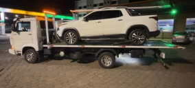 Guincho plataforma em Betim transportando um veículo branco, estacionado em um posto de combustíveis à noite.