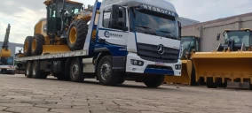 Guincho plataforma transportando uma retroescavadeira amarela em Campo Grande, com outros equipamentos visíveis ao fundo.