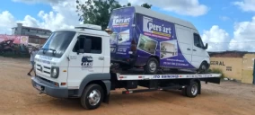 Guincho plataforma transportando uma van branca com adesivo, estacionado em Valença, em área de terra.