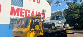 Guincho plataforma amarelo transportando um carro branco em Campo Mourão, estacionado em frente a uma oficina mecânica.