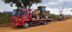 Guincho plataforma vermelho transportando uma retroescavadeira amarela em Córrego Danta, estacionado em área de terra.