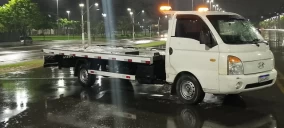 Guincho plataforma branco estacionado em Hortolândia, com luzes de emergência acesas, em área molhada.