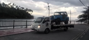 Guincho plataforma em Hortolândia transportando um veículo azul, estacionado próximo a uma área com vegetação e água.